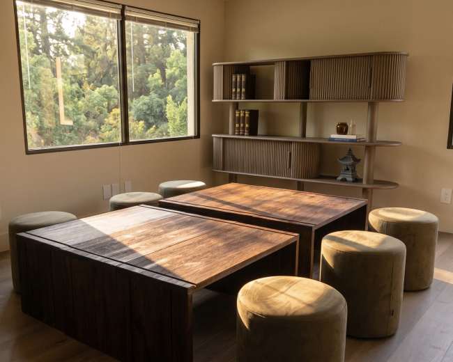 The image shows a minimalist room with two wooden tables, cylindrical stools, and large windows overlooking a green landscape.