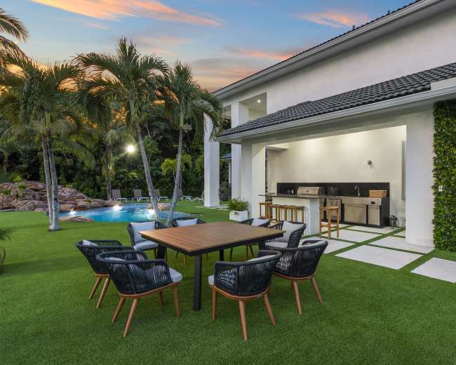 A modern outdoor dining area features a wooden table surrounded by chairs, adjacent to a pool and a kitchen setup in a lush green yard.