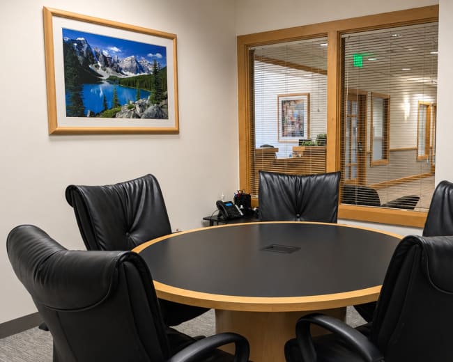 The image shows a conference room with a round table surrounded by four black leather chairs and a window overlooking a hallway.