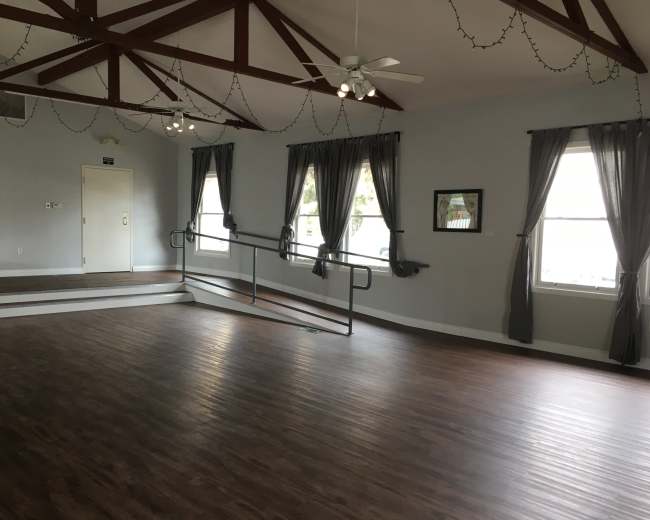 The image shows a spacious, empty room with wooden flooring, high ceilings supported by exposed beams, and large windows dressed with dark curtains.