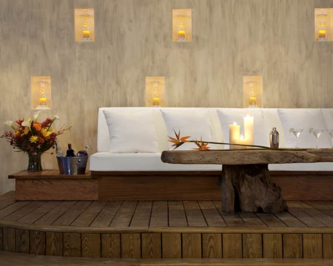 A wooden table with decorative items sits in front of a white couch against a textured wall, illuminated by candles placed in wall sconces.
