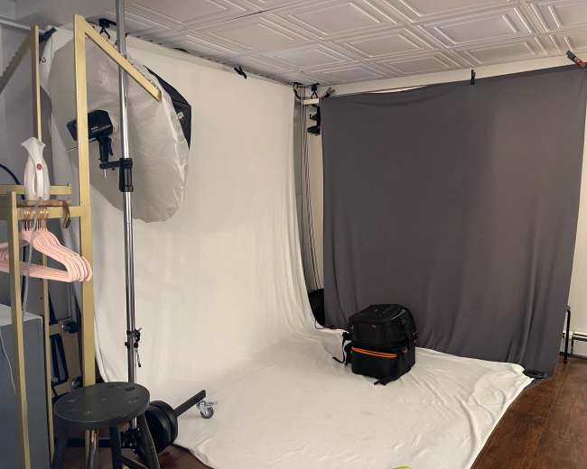 The image shows a photography studio setup with a gray backdrop, a white backdrop, a lighting umbrella, a stool, and a bag on the floor.