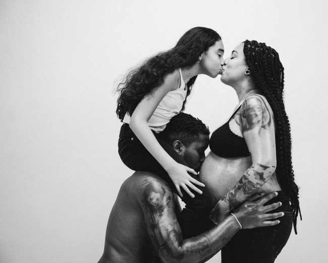 A pregnant woman and her partner are positioned against a white background, while a girl kisses the woman's cheek, sitting on the partner's shoulders.