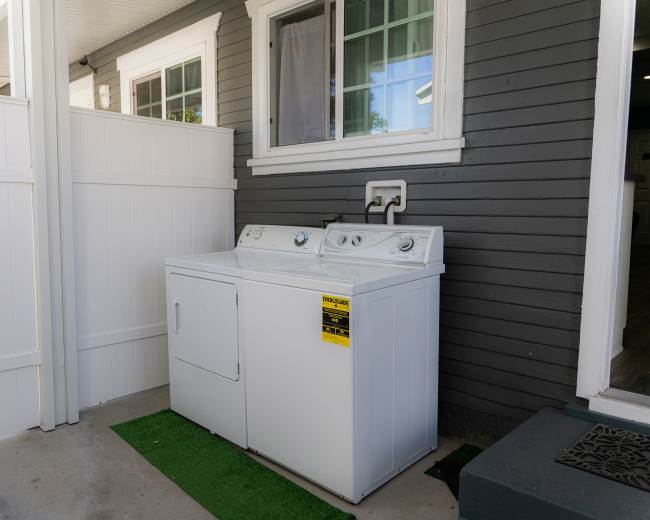 A white washing machine and dryer set are placed against a gray house wall, beside a fenced area.