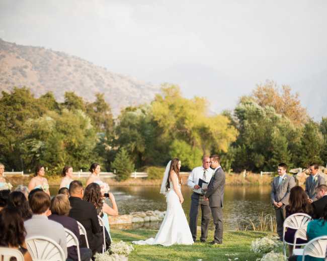 A bride and groom exchange vows during an outdoor wedding ceremony by a pond, surrounded by guests and a scenic mountainous backdrop.