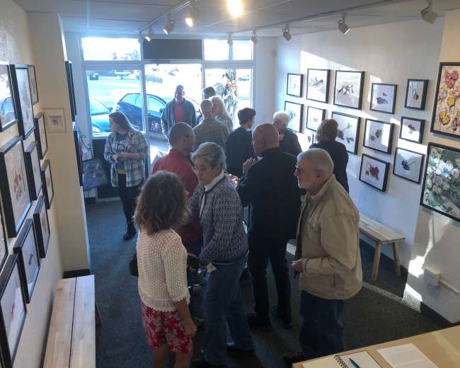 A group of people stands inside an art gallery, surrounded by framed artworks on the walls, with some conversing and others observing the pieces.