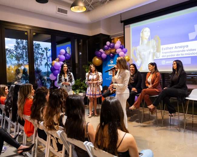 A group of women sits on a stage while another woman speaks to an audience during an event.