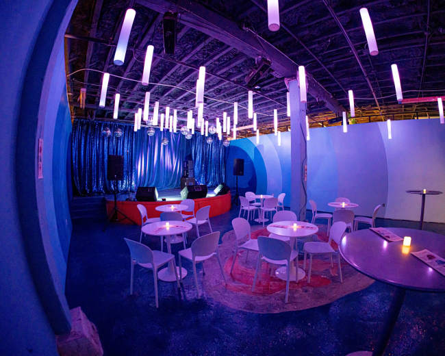 The image shows a venue with blue walls, small white tables and chairs, and a stage area with hanging lights.