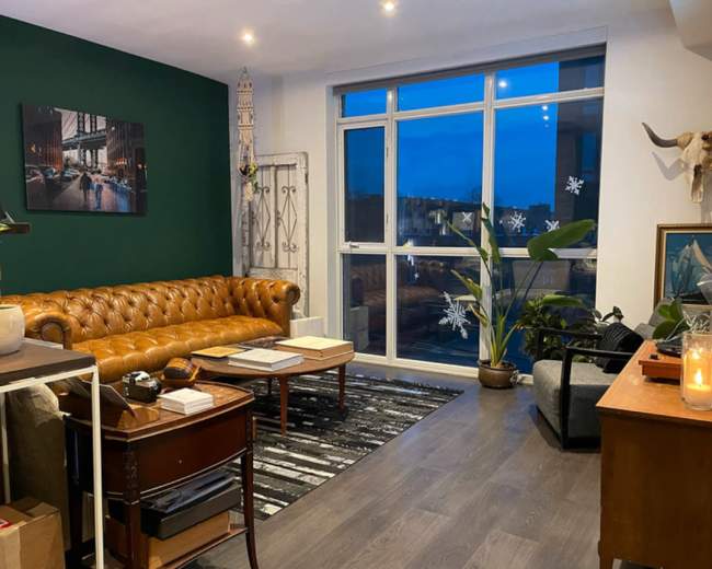 The image shows a cozy living room with a brown leather couch, a coffee table, a variety of plants, and large windows allowing natural light to enter.