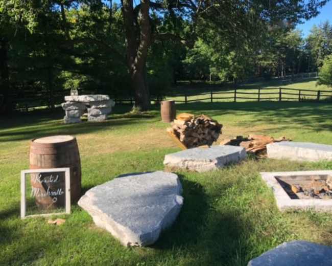 The image shows an outdoor setting with stacked firewood, a stone fire pit, large stones, a barrel, and a sign that says "Welcome to Marshmellow."