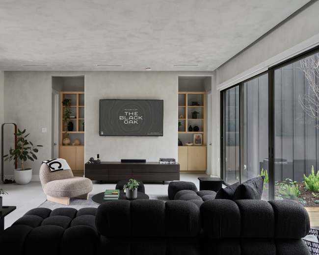 The image shows a modern living room with a black sectional sofa, a round coffee table, and a flat-screen TV mounted on the wall above built-in shelves.