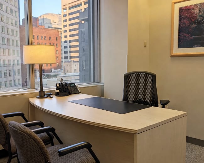 An office space features a desk, a lamp, and two chairs, with large windows showcasing a view of city buildings.