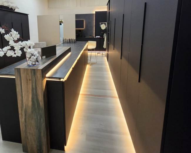 The image shows a modern interior space featuring sleek black cabinetry with a wooden accent along a well-lit corridor.