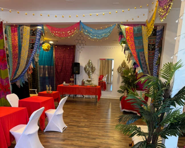 The image shows a decorated event space with red tables, colorful fabric drapes hanging from the ceiling, and a few potted plants.