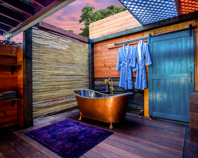 The image shows a wooden outdoor bathroom featuring a copper bathtub, two blue robes hanging on a wall, and a statue of Buddha on a side table.