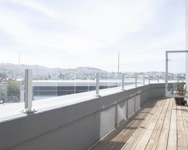 A spacious balcony with a wooden floor and a glass railing, overlooking a cityscape under a clear sky.