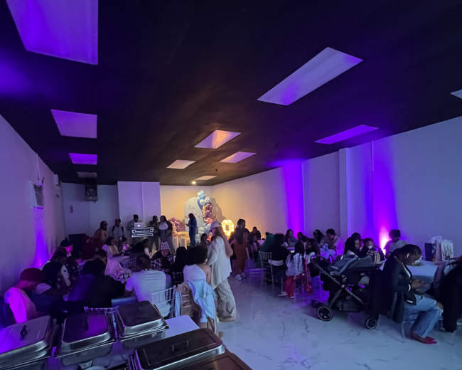 A dimly lit room filled with people seated at tables, some standing, with food trays arranged along the side.
