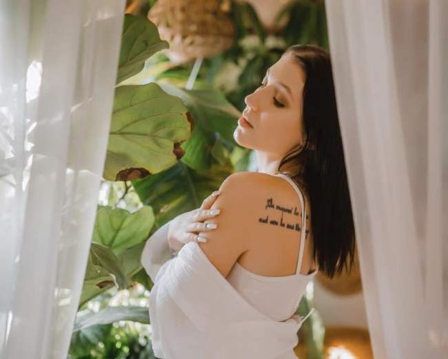 A woman in a white outfit poses beside lush green plants, with sheer curtains framing the scene.