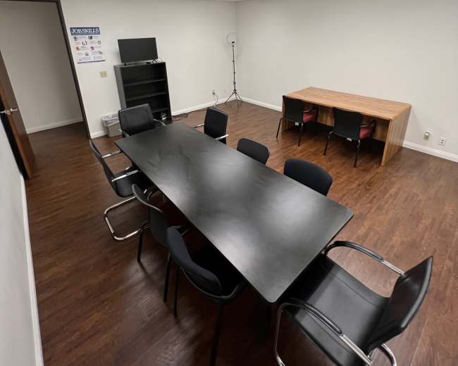 A modern office meeting room featuring a long black conference table surrounded by chairs, with a bookshelf and a wooden table in a well-lit space.