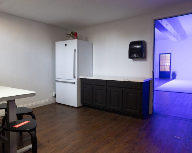 The image shows a tidy room with a white refrigerator, a dark cabinet, a table with chairs, and a doorway leading to another brightly lit space.