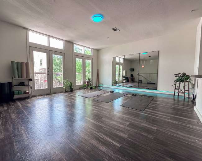 The image shows a spacious, bright room with large windows, hardwood flooring, and yoga mats laid out, alongside yoga props stacked against the wall.