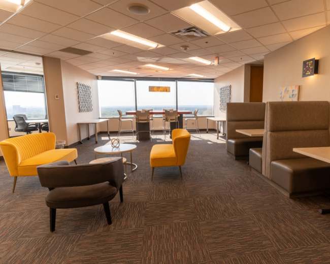 The image shows a modern office retreat with various seating arrangements, including colorful chairs and booths, and large windows overlooking a landscape.