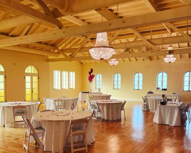 A spacious event hall with several round tables covered in white tablecloths, adorned with sparkling chandeliers and large windows letting in natural light.
