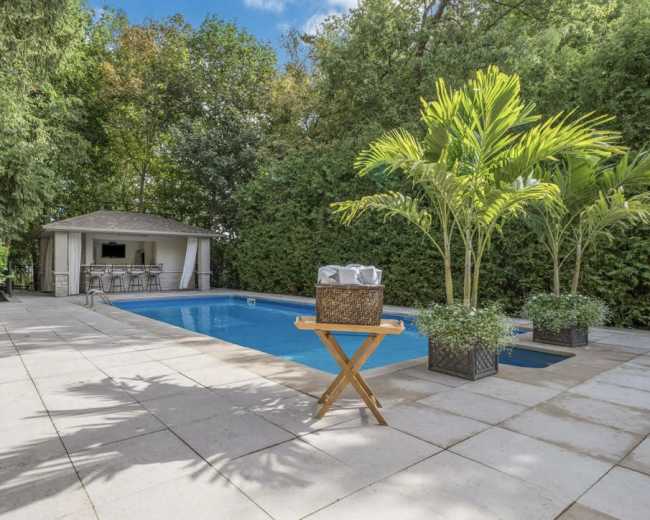 A rectangular swimming pool is surrounded by tiled flooring, lush greenery, and features a gazebo with seating in the background.