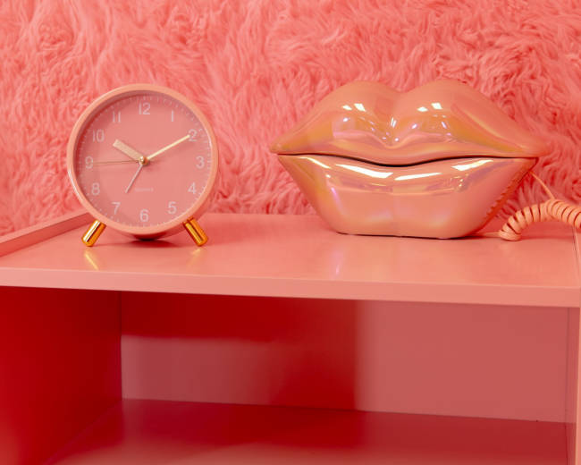 A pink shelf features a round clock and a ceramic lip-shaped decor piece against a textured pink background.