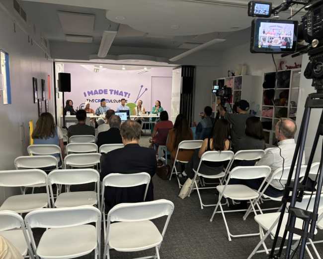 A panel discussion is taking place in a room with a seated audience, featuring four speakers at a table in front of a backdrop that reads "I MADE THAT!"