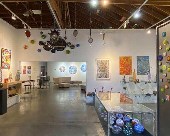 The image shows an art gallery featuring a variety of colorful glass artworks displayed on walls and shelves, with a seating area visible in the background.