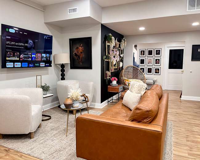 The room features a leather sofa, two white chairs, a round table, and a large television on the wall, with framed artwork and decorative elements throughout.