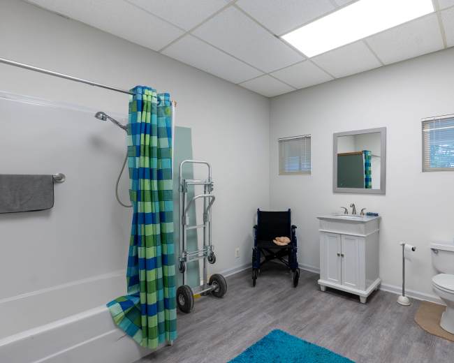 The image shows a bathroom equipped with a bathtub, a wheelchair, a sink unit, and a toilet, featuring a blue and green shower curtain.