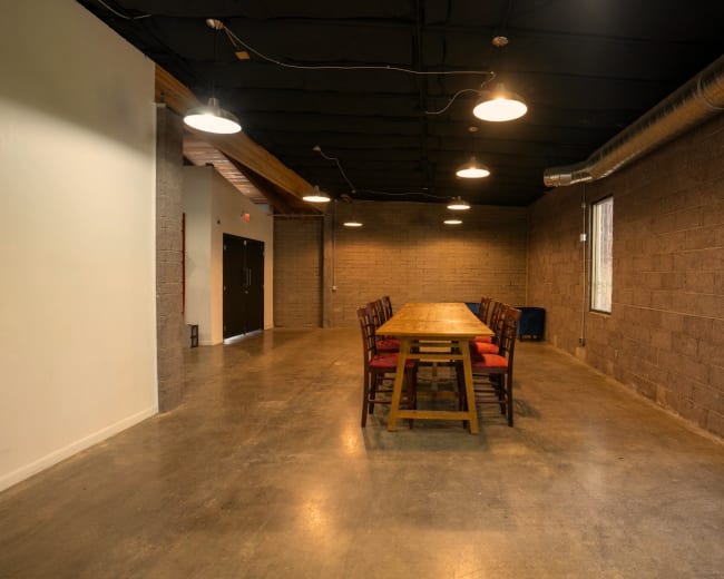 A spacious room features a long wooden dining table with chairs arranged around it, flanked by concrete walls and illuminated by overhead lighting.