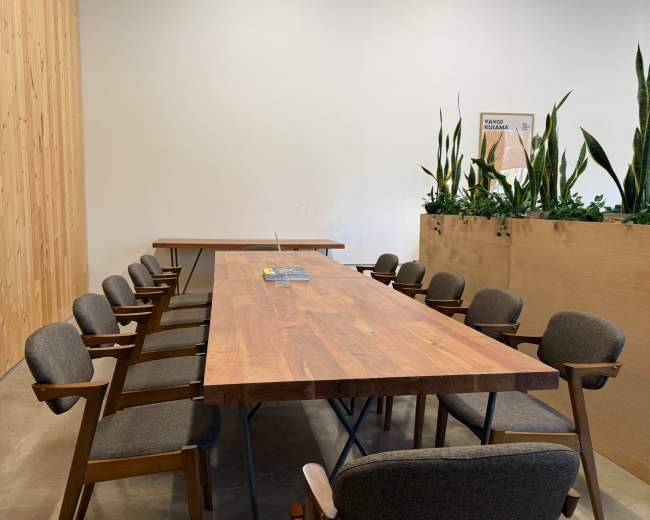 A long wooden conference table with several gray chairs is positioned in a well-lit, minimalist room featuring a planter with tall green plants.