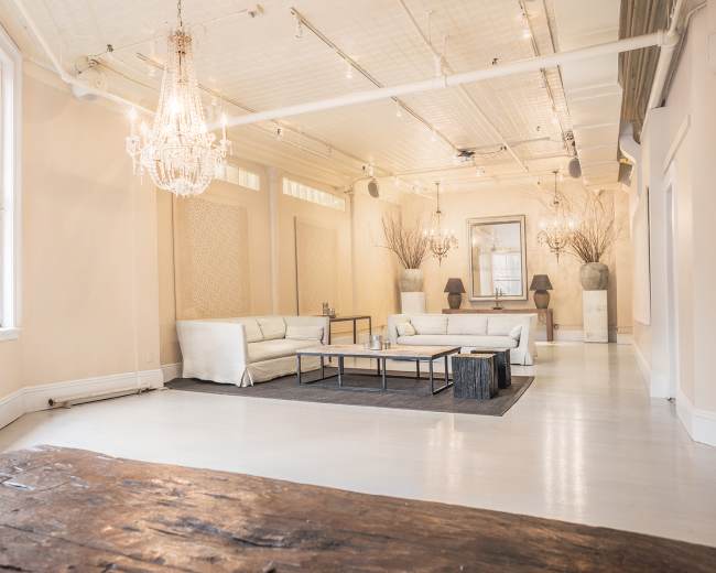 The image shows a spacious interior with light-colored walls, a central seating area featuring a white sofa set, a dark area rug, and elegant lighting fixtures.