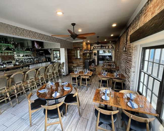 The image shows a restaurant interior featuring wooden tables set with blue napkins, surrounded by chairs, and a bar area with shelves of bottles in the background.