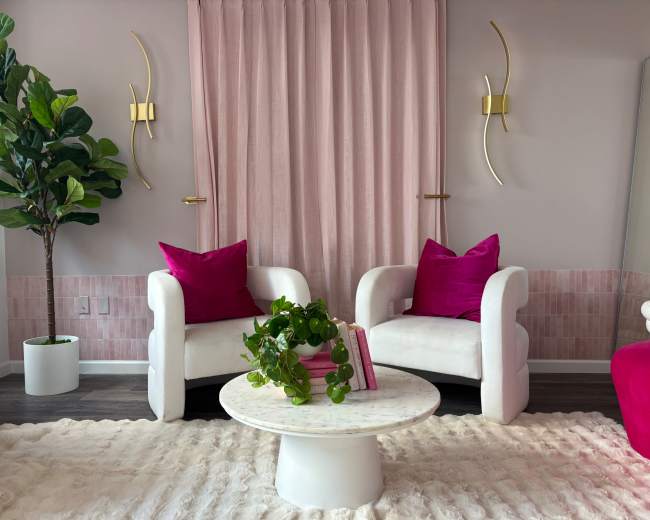 The scene features two white armchairs with pink cushions, a round marble coffee table, a potted plant, and pink curtains in a bright room.
