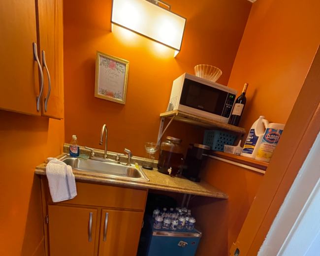 The image shows a small kitchenette area featuring a sink, a microwave, and storage cabinets, with various cleaning supplies and bottled water on display.