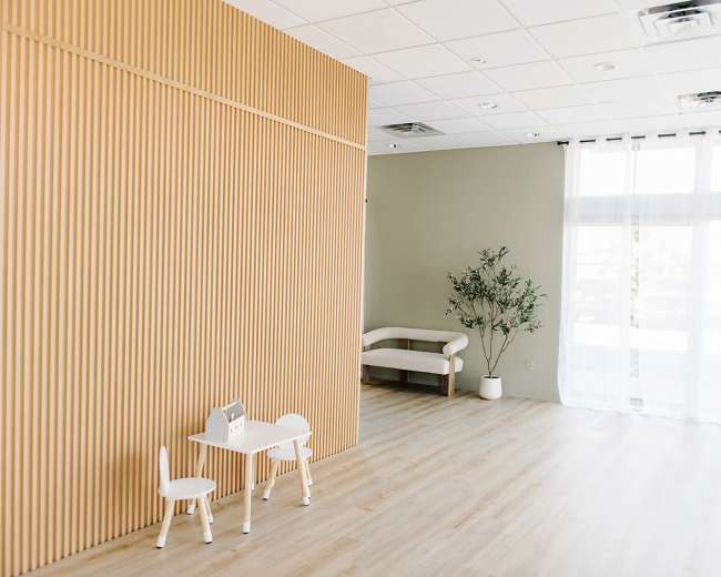 The room features a wooden-paneled wall, a small table with two chairs, a couch, and a potted plant beside large windows covered with sheer curtains.