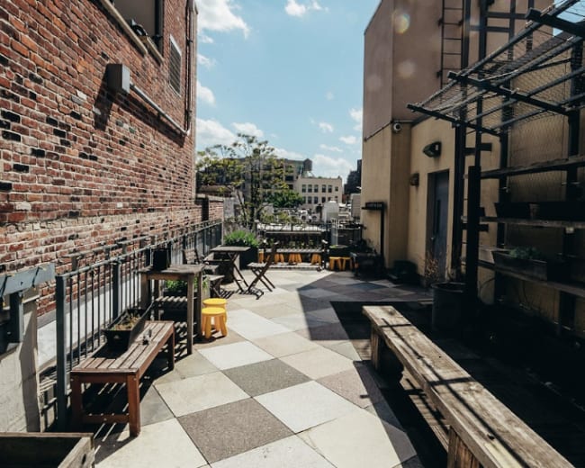 The image shows a rooftop terrace with a patterned stone floor, wooden benches, and planters under a bright blue sky with a few clouds.
