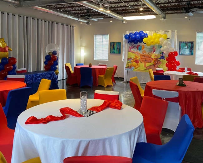 The image shows a decorated party room with tables covered in white tablecloths and adorned with colorful chairs and balloons.
