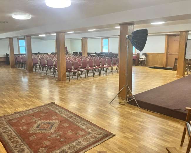 The image shows a spacious room with wooden floors, arranged seating with pink chairs facing a small stage, and a large light fixture overhead.