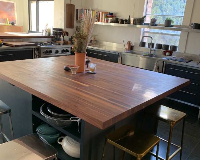 The image shows a modern kitchen with a large wooden island, surrounded by metal stools, and shelves filled with various kitchenware.