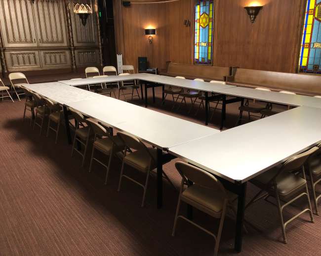 A large meeting room features an arrangement of tables in a U-shape, surrounded by folding chairs, with stained glass windows in the background.