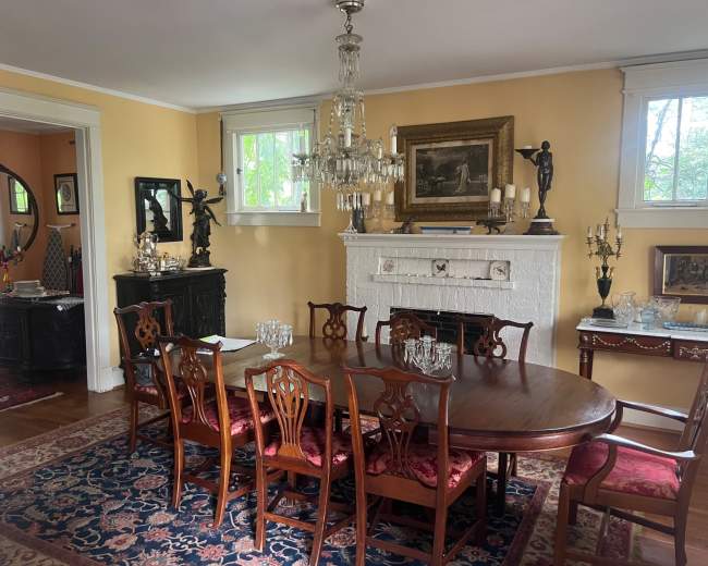 The image shows a dining room with a wooden table surrounded by chairs, a chandelier, artworks on the walls, and a fireplace adorned with decorative items.
