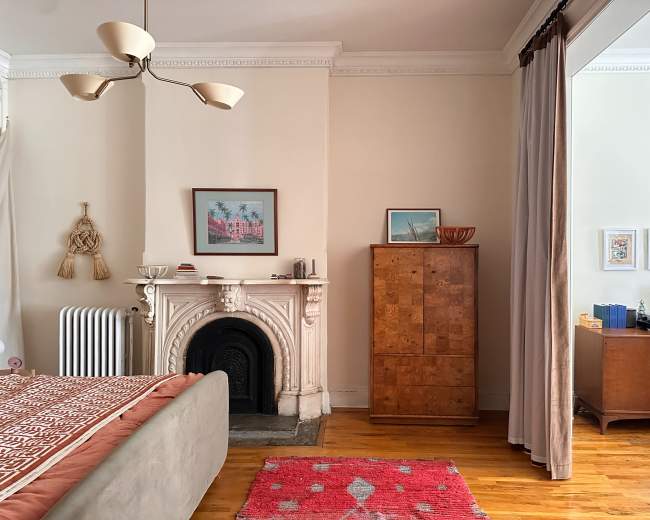 The image shows a cozy bedroom featuring a decorative fireplace, wooden furniture, and a pink rug on a hardwood floor.