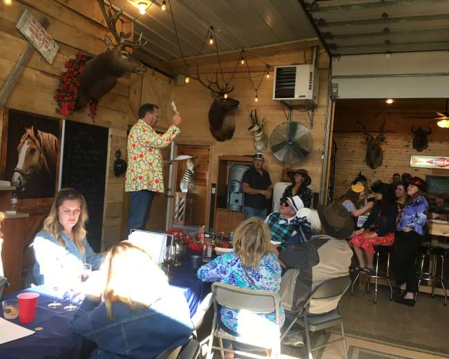 A man in a brightly patterned shirt stands at a podium, while a diverse group of people socializes in a rustic gathering space adorned with taxidermy and wooden walls.
