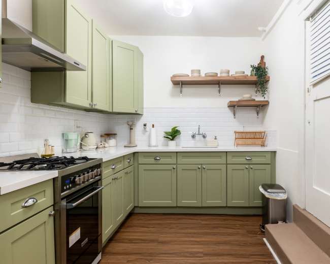 A compact kitchen features green cabinetry, a stainless steel gas stove, and wooden shelves with various kitchen items.