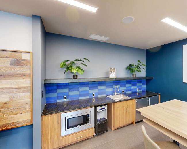 The image features a modern kitchen space with a blue tiled backsplash, a stainless steel sink, a microwave, and a wooden dining table.